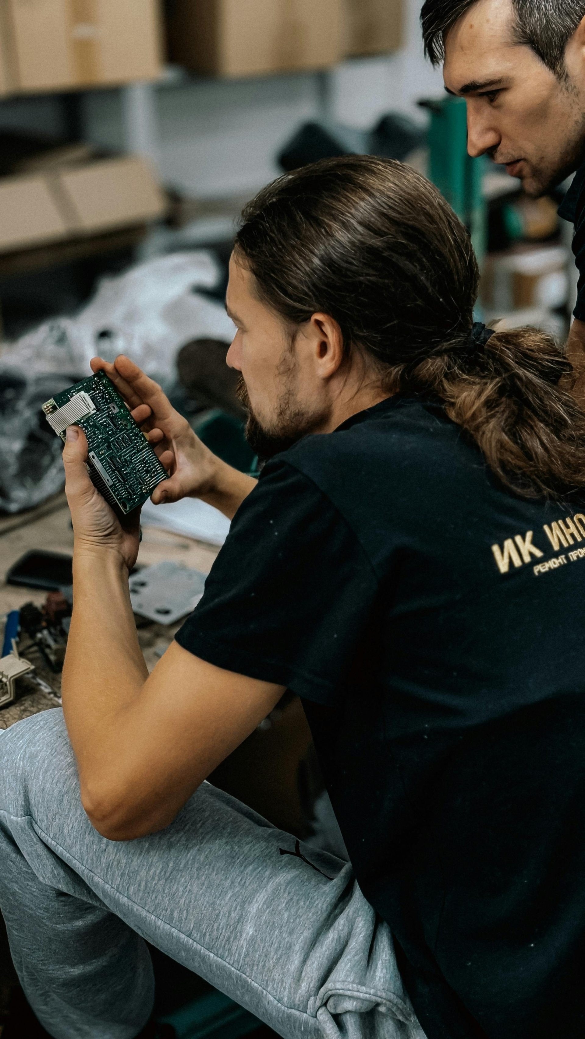 Two technicians carefully examining a circuit board in a cluttered workshop environment.